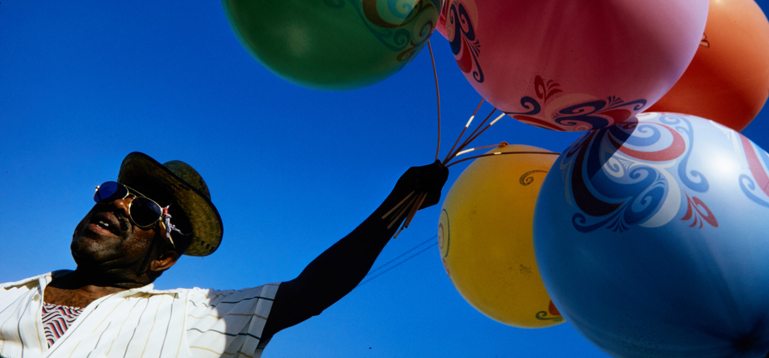 A man in sunglasses and a straw hat holds a bunch of balloons against a blue sky, Chicago, 1970s